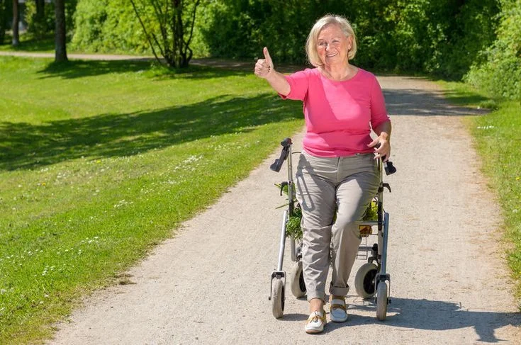 Senior woman in pink walking on a path with a rollator giving a thumbs up